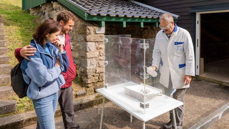 Eine Gruppe von drei Personen, darunter ein Mann in einem weißen Kittel, steht um einen Tisch mit einem Experiment. Die beiden Zuschauer schauen gespannt zu, während der Experte etwas in den Behälter gießt.