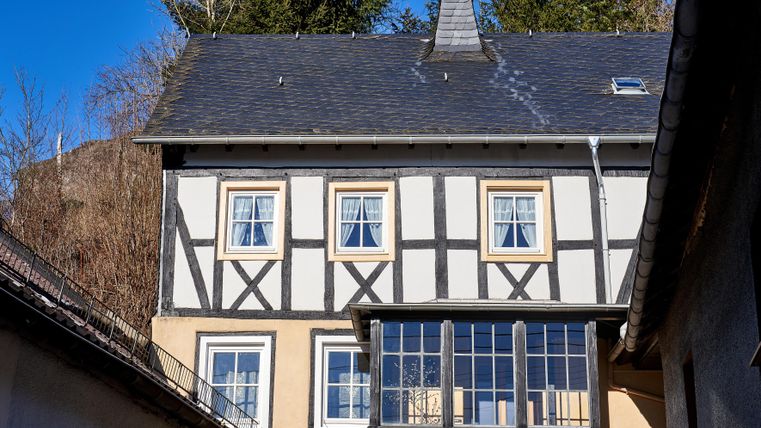 A traditional half-timbered house with white walls and dark wooden frames. In the foreground, you can see windows overlooking the surrounding trees.