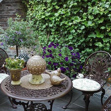 Ein gemütlicher Garten mit einem runden Tisch aus Metall und geschwungenen Stühlen. Umgeben von grünen Pflanzen und bunten Blumen, strahlt der Bereich eine entspannte Atmosphäre aus.