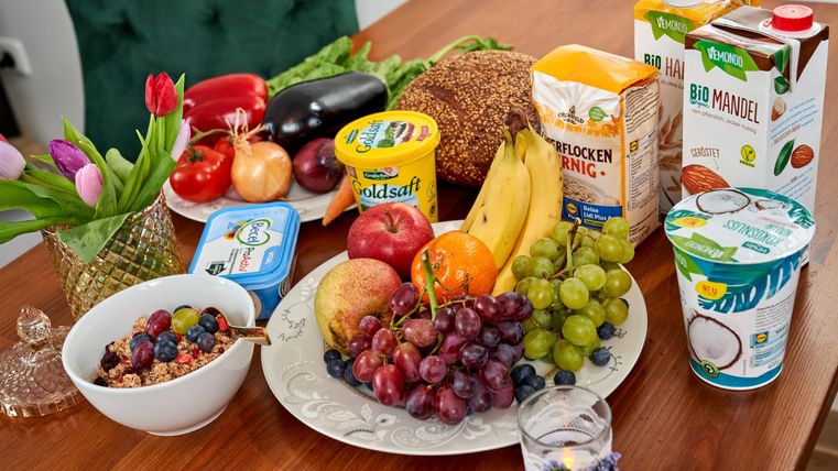 A table with a colorful selection of fresh fruits, including grapes, apples, and bananas. Next to the fruit, various drinks and snacks are prepared.