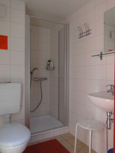 Ein einfaches Badezimmer mit einer Dusche, einem Waschbecken und einer Toilette. Die Wände sind gefliest und die Ausstattung ist funktional.