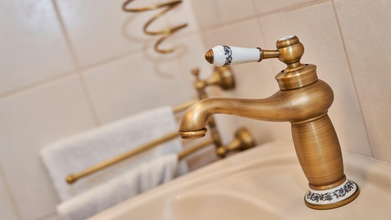 An elegant faucet made of golden metal in a bathroom. In the background, towels and a decorative rod are visible.