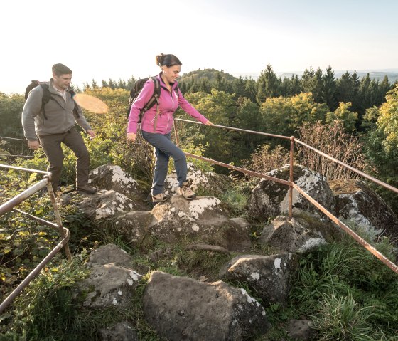 Zwei Wanderer auf einem felsigen Pfad mit Geländer, umgeben von Wald und Aussicht auf Hügel., © Eifel Tourismus GmbH, Dominik Ketz
