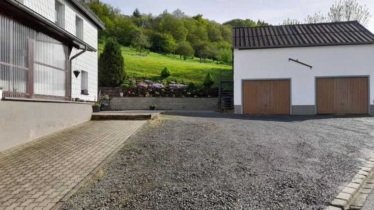 A quiet driveway with a house on the left side and a garage on the right side. In the background, green hills and trees are visible.