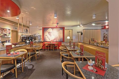 Cozy café with wooden furniture, tables, and chairs. On the wall, a large picture of a baker. Red accents and menus on the tables.
