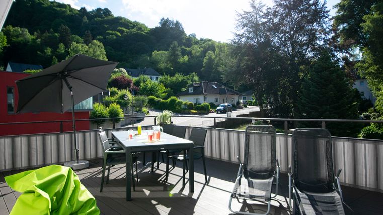 Eine sonnige Terrasse mit einem Tisch und Stühlen. Im Hintergrund sind grüne Hügel und Wohnhäuser zu sehen.