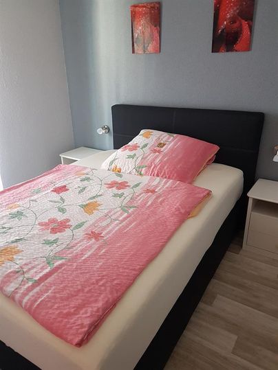 Ein gemütliches Schlafzimmer mit einem Doppelbett und bunten Kissen. An der Wand hängen zwei Bilder, und es gibt einen Nachttisch neben dem Bett.