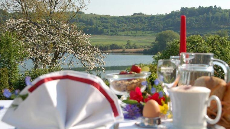 Ein schöner Frühstückstisch im Freien mit Blick auf einen See und grüne Hügel. Der Tisch ist mit frischen Speisen und einer Blume dekoriert.