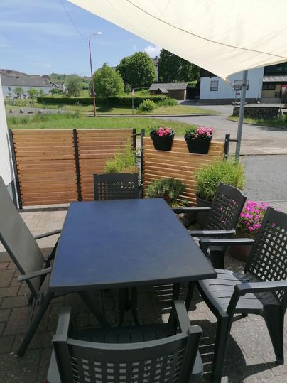 Een gezellige buitenruimte met een zwarte tafel en stoelen. De ruimte is versierd met planten en bloeiende bakken.