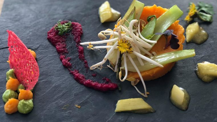 Ein kunstvoll angerichtetes Gericht auf einem schwarzen Teller. Es enthält verschiedene Gemüsesorten, Saucen und essbare Blumen.