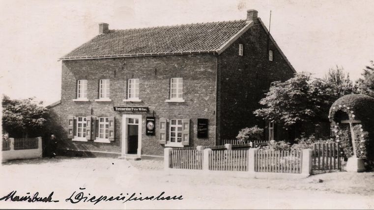 Ein altes, gemauertes Haus mit weißen Fenstern und einem Bauernhof-ähnlichen Charme. Vor dem Haus sind Bäume und ein Garten mit einem Zaun zu sehen.