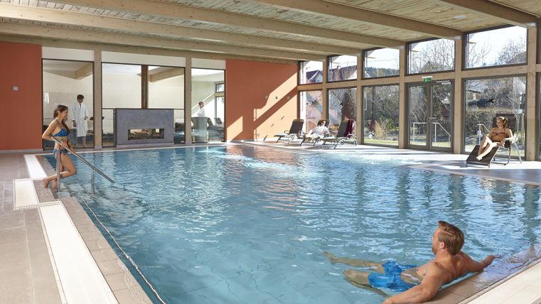 Ein modernes Hallenbad mit großen Fenstern und natürlichem Licht. Menschen entspannen am Beckenrand und im Wasser.