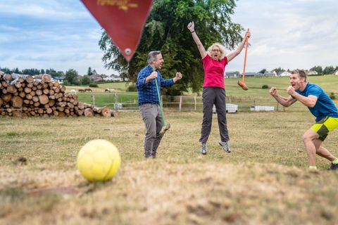 Drie personen hebben plezier op een grasveld. Ze vieren en springen van blijdschap, terwijl een gele bal op de voorgrond ligt.