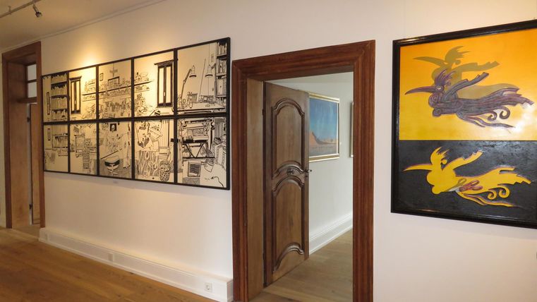 Ein heller Innenraum mit Holzfußboden, der eine Galerie mit verschiedenen Kunstwerken an der Wand zeigt. Durch eine Tür ist ein weiterer Raum sichtbar.