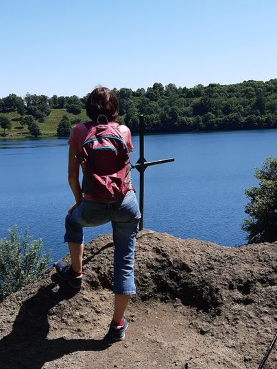 Een persoon zit op een rots met uitzicht op een rustige vijver. De zon schijnt en de omgeving is groen en bosrijk.