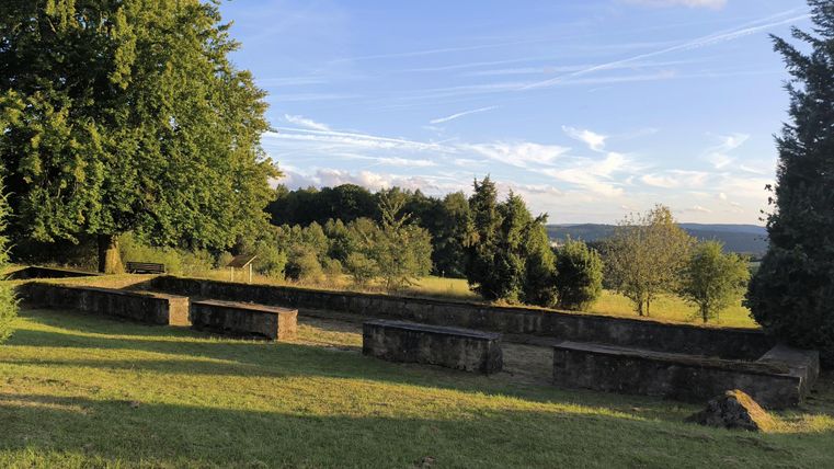 Ein ruhiger Friedhof mit alten Grabsteinen, umgeben von Bäumen und Wiesen. Der Himmel ist blau mit einigen Wolkenstreifen.