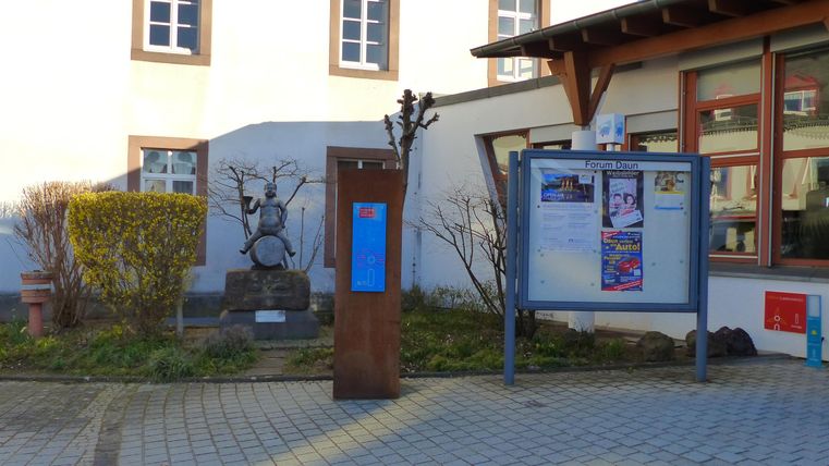 Ein Platz vor einem Gebäude mit einer Informationswand und einem Statue. Der Bereich ist ordentlich mit Grünflächen und gepflastertem Boden.