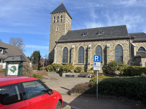 Eine alte Kirche mit einem markanten Turm steht im Vordergrund. Im Vordergrund ist ein rotes Auto und rundherum sind gepflegte Pflanzen und eine Parkzone zu sehen.