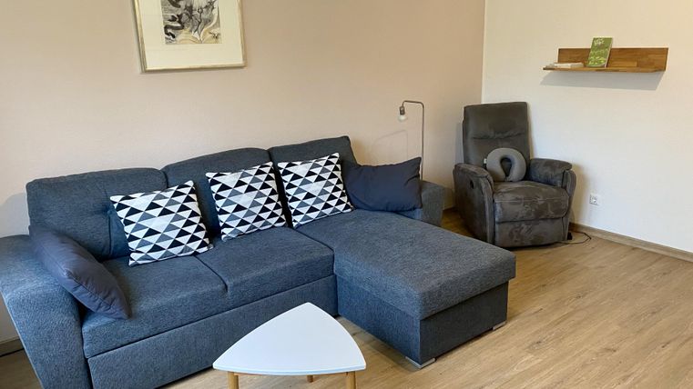 Ein modernes Wohnzimmer mit einem grauen Sofa und stilvollen Kissen. Im Hintergrund befinden sich ein Sessel und eine kleine Ablage.