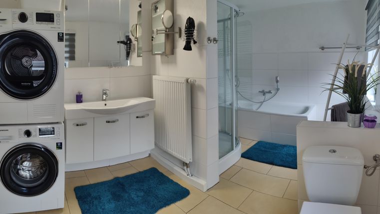 A modern bathroom with a washing machine and dryer, a sink, and a shower. There are blue mats and a windowsill with plants.
