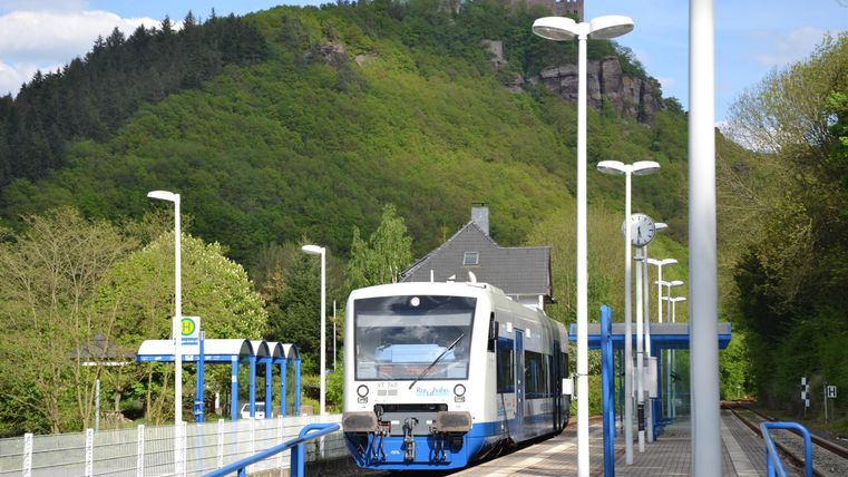 A train is waiting at a station, surrounded by green hills. The sun is shining and it is a clear day.