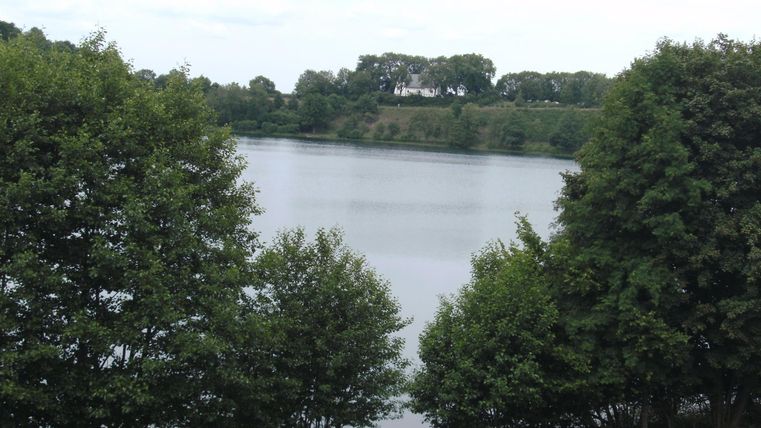 Ein ruhiger See, umgeben von grünen Bäumen. Im Hintergrund sind einige Menschen am Ufer sichtbar.