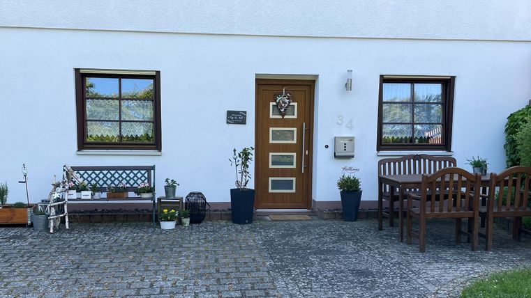Een uitnodigende huisingang met een houten deur en twee ramen. Voor de deur staan bloempotten en een bankje.