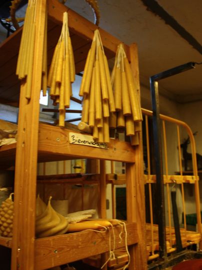 Ein Holzregal mit mehreren langen, gelben Kerzen. Im Hintergrund sind weitere Bastelmaterialien und Werkzeuge zu sehen.