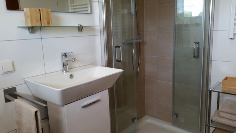 Ein modernes Badezimmer mit einer Dusche und einem Waschbecken. Die Wände sind hell gefliest und es gibt eine kleine Ablage.