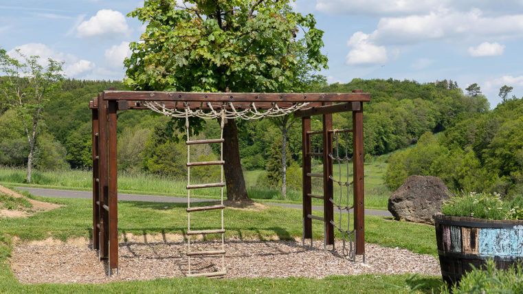 Een speelplaats met een klimtoestel van hout en touwen. Op de achtergrond zijn groene bomen en een blauwe lucht zichtbaar.