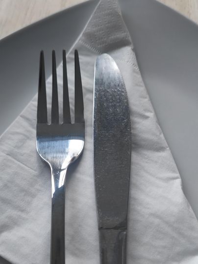 Ein Gedeck mit einer Gabel und einem Messer auf einem weißen Servietten. Im Hintergrund ist ein grauer Teller zu sehen.