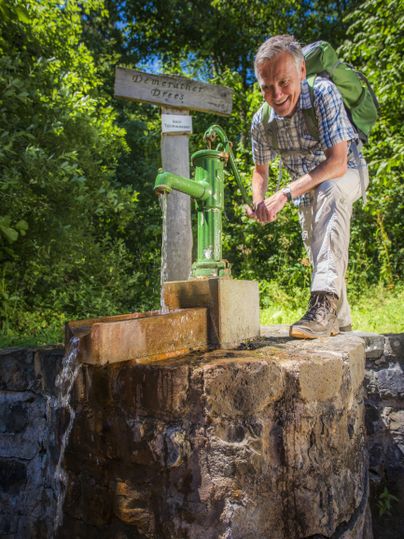 Ein Mann steht an einer Handpumpe im Freien. Um ihn herum sind Bäume und ein Wegweiser sichtbar.