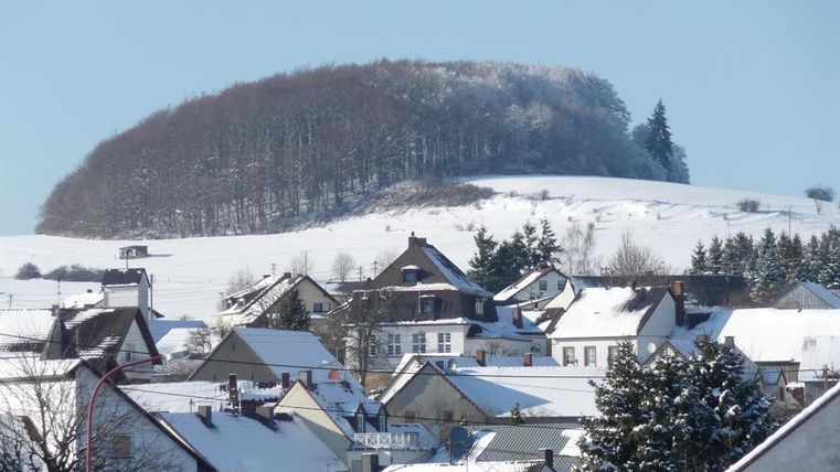 Een met sneeuw bedekt landschap met een heuvel op de achtergrond en een klein dorp op de voorgrond. De daken van de huizen zijn bedekt met sneeuw en de bomen op de heuvel zijn winterachtig besneeuwd.