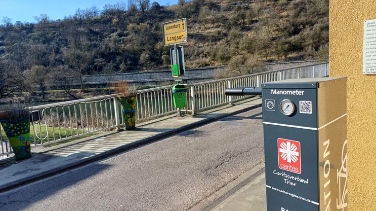 Eine Fahrrad-Service-Station mit einem Manometer steht an einer ruhigen Straße. Im Hintergrund ist eine grüne Landschaft und ein Schild mit dem Ortsnamen sichtbar.