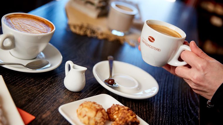 Eine Tasse Kaffee wird gehalten, während auf dem Tisch Kekse und eine weitere Tasse stehen. Der Tisch hat eine dunkle Holzoberfläche.