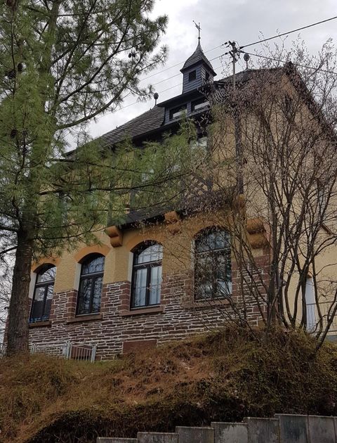 Ein historisches Gebäude mit verzierten Fenstern und einem kleinen Turm. Es liegt auf einem Hang, umgeben von Bäumen und trockenen Sträuchern.