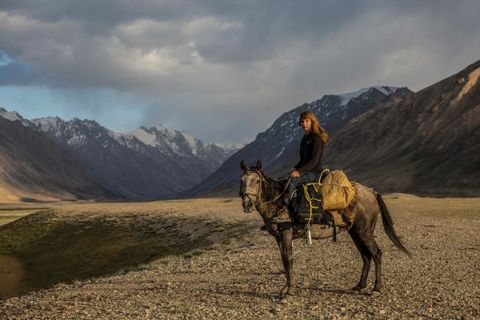 A rider sits on a gray horse in a mountainous landscape. Majestic mountains and a dramatic sky surround her.
