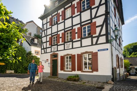 Een charmant vakwerkhuis met witte muren en rode luiken. Twee personen wandelen rustig langs de straat.