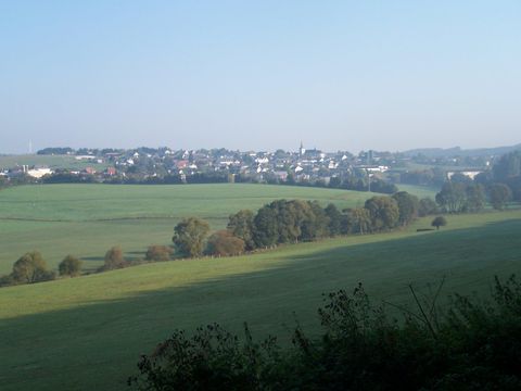 A picturesque landscape with gentle hills and a small town in the background. The sky is clear and the meadows are green.