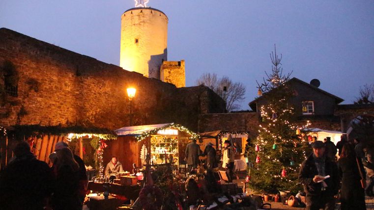 Een kerstmarkt met feestelijk versierde kraampjes en een grote kerstboom. Op de achtergrond staat een verlichte toren onder een heldere avondhemel.