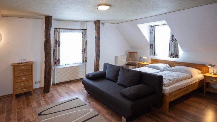 Ein gemütliches Schlafzimmer mit Holzboden und hellem Licht. Es gibt ein großes Bett, ein Sofa und Fenster mit Vorhängen.