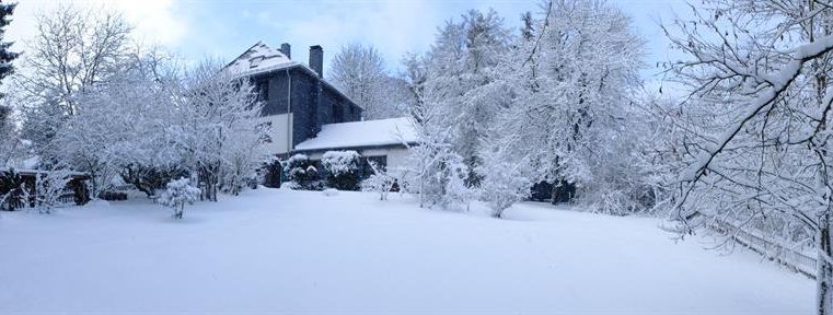 Een winterlandschap met een met sneeuw bedekt terrein en bomen. Op de achtergrond staat een gezellig huis onder een heldere lucht.