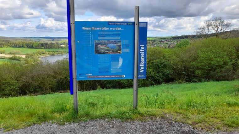 An information sign on a slope overlooking a green landscape and a lake. The sky is overcast and the surroundings are calm and close to nature.