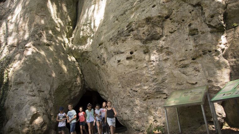 Eine Gruppe von Menschen steht vor einer großen Felsformation und einer Höhle. Die Umgebung ist grün und natürlich.