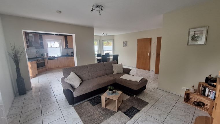 Ein modernes Wohnzimmer mit einem grauen Sofa und einem kleinen Tisch. Im Hintergrund sieht man eine offene Küche und einen Essbereich.