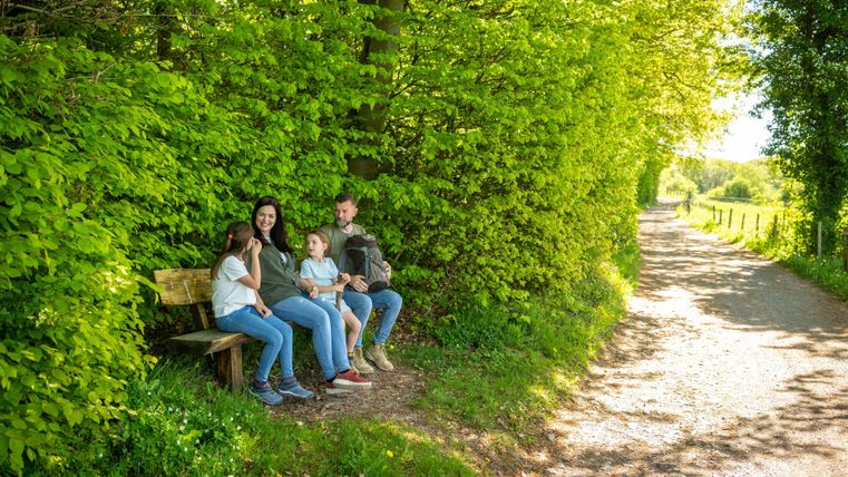 Een gezin zit op een bank naast een schaduwrijke weg. De weg is omgeven door weelderig groen en bomen.