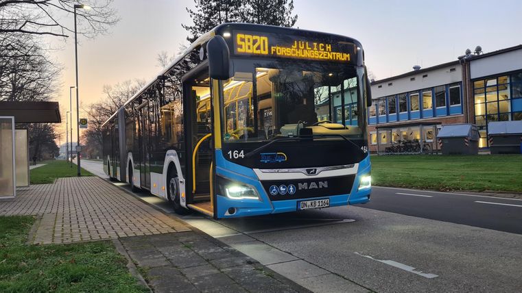 A modern bus stops at a station. Buildings and trees are visible in the background.