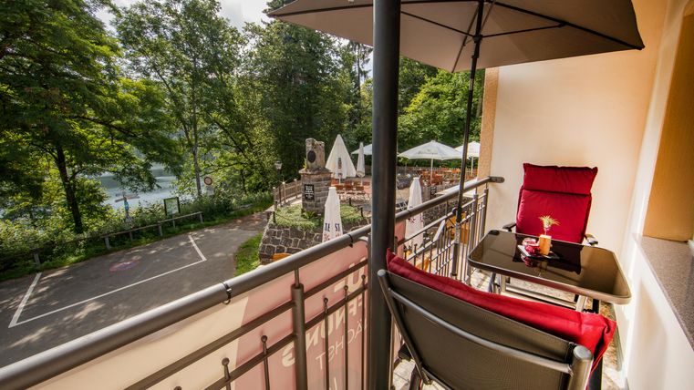Ein komfortabler Balkon mit einem Tisch und Stühlen, umgeben von Bäumen. Im Hintergrund sieht man einen Gartenbereich mit Sonnenschirmen.