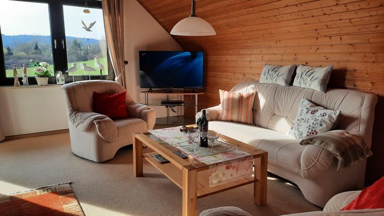 A cozy living room with wooden paneling and large windows. There are comfortable sofas, a coffee table, and a television.