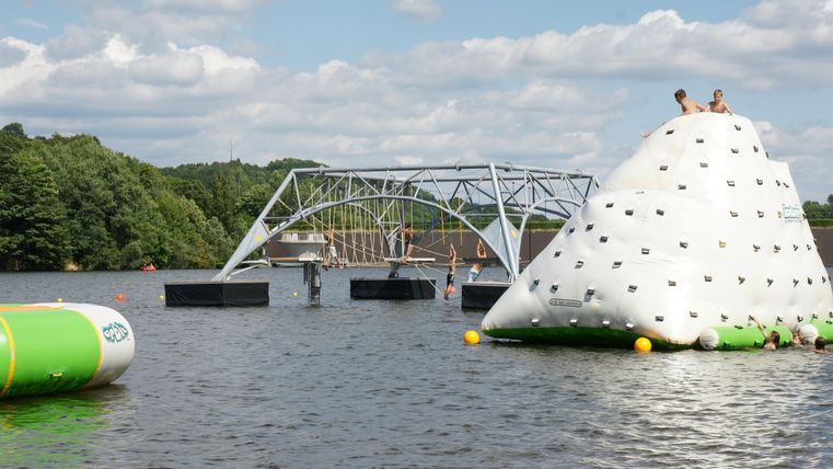 Ein Wasserspielplatz mit einer Kletterwand und einer großen aufblasbaren Fläche. Im Hintergrund ist eine Brücke und grüne Bäume zu sehen.